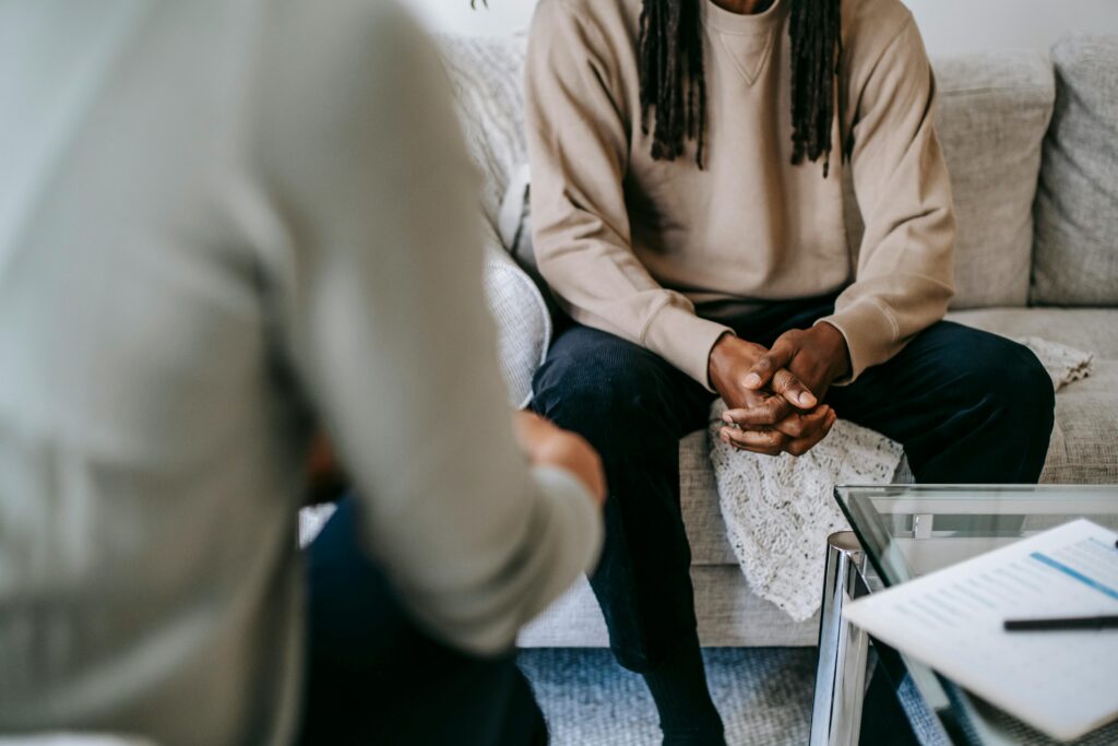 Photo of two people sitting down across from each other in a therapy session. There is a man with black slacks and a beige sweater with his hands clasped together.
